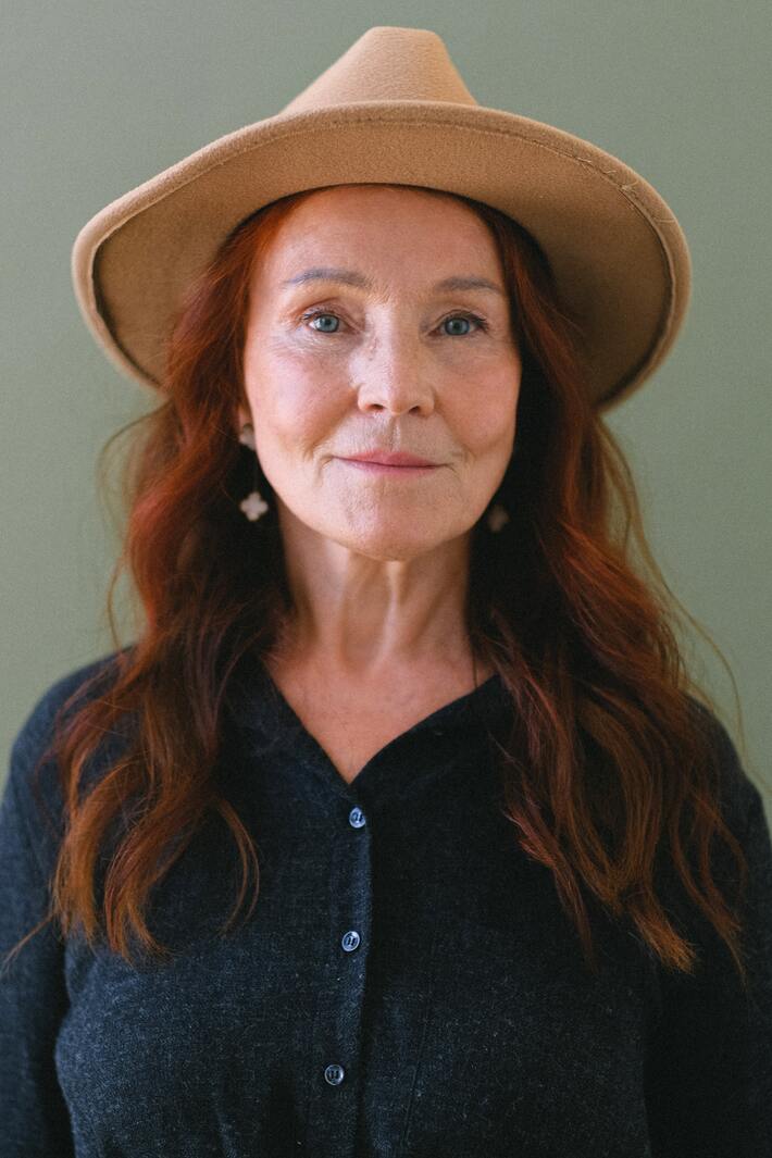 A redheaded women in her 50s in a wide brim hat, looks at the camera with a small smile.