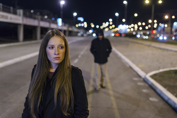 A woman walks down a dark street with a man in a black hoodie following behind her